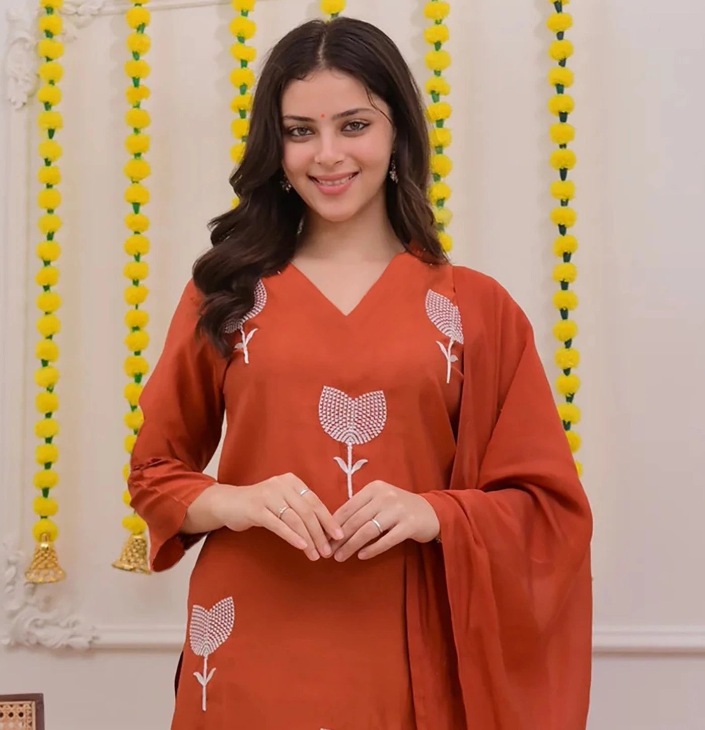 Woman wearing an orange traditional outfit with floral patterns against a decorative background.
